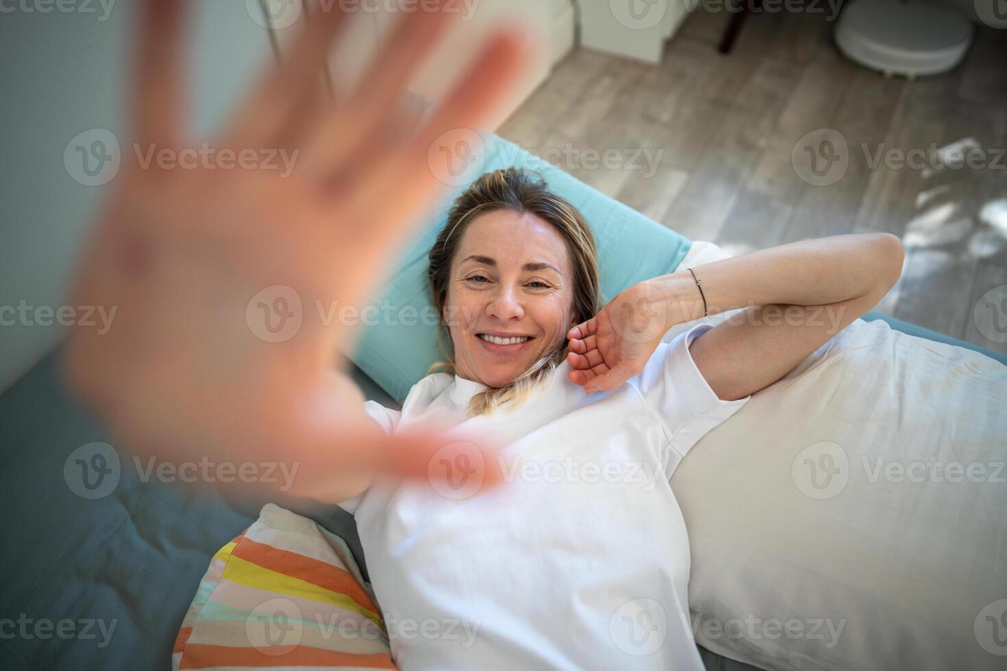 Sleepy middle-aged woman lies on bed smiling and stretching raised one hand close up to camera. photo