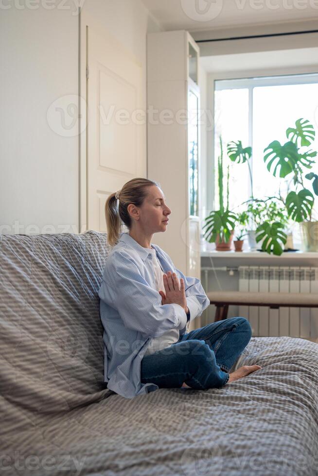 Woman sits on couch with eyes closed in lotus pose doing meditation practices, breathing exercises. photo