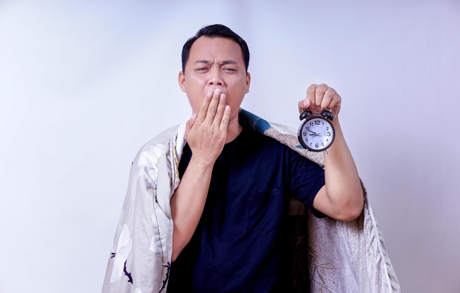 A tired asian man yawns while holding a clock, wrapped in a blanket, conveying morning fatigue. photo