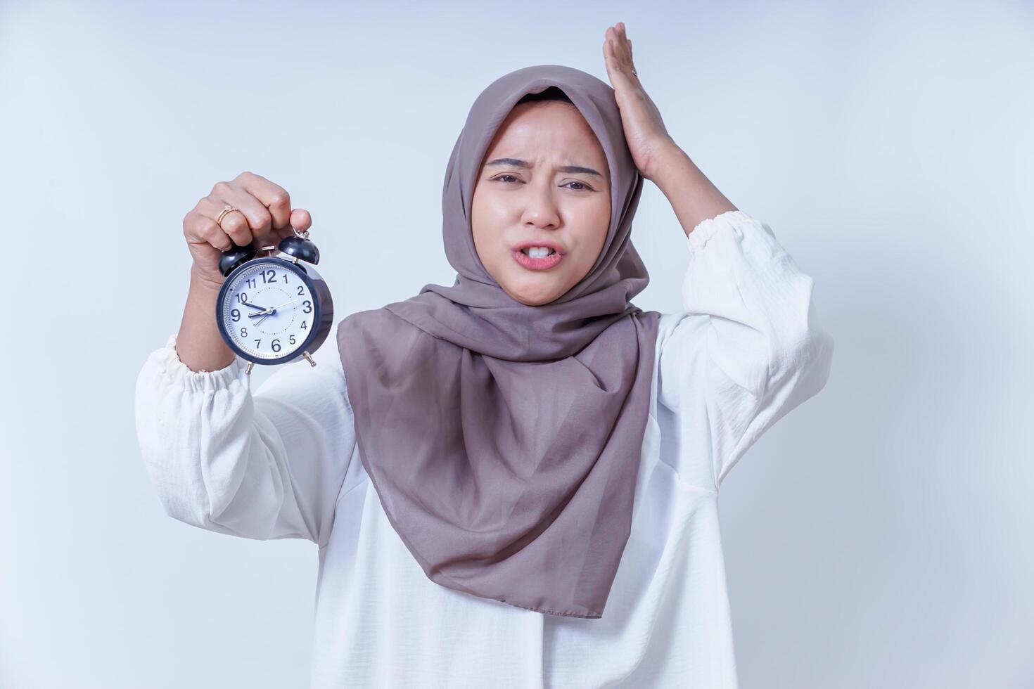 Frustrated asian woman in a hijab holding a clock, showing urgency and concern about time in a white background. photo