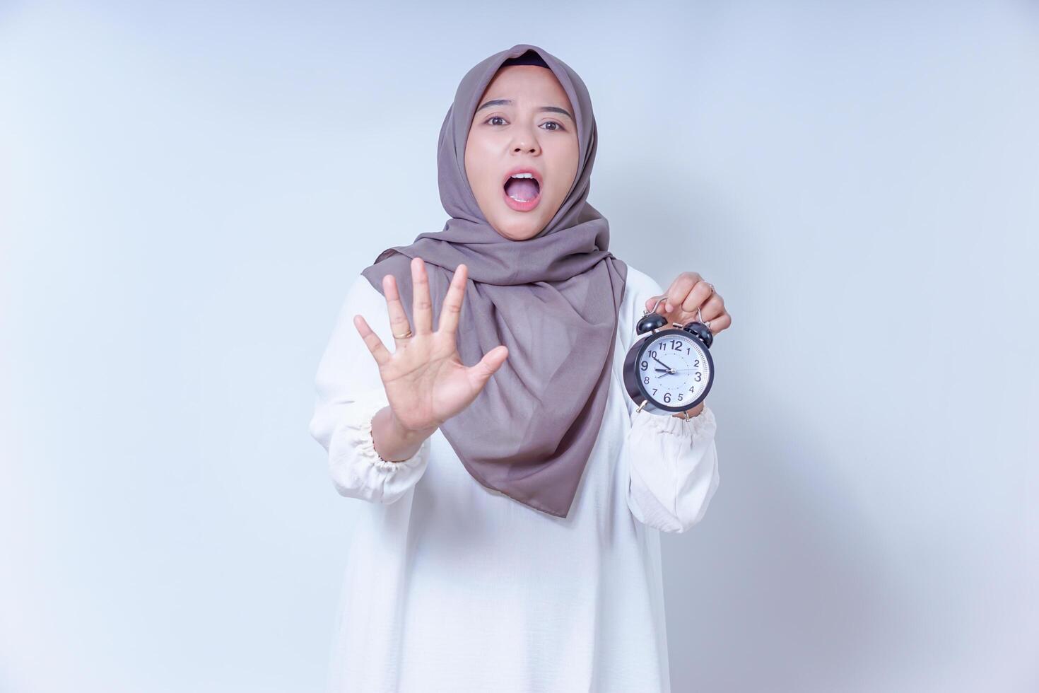 A surprised asian muslim woman holds an alarm clock, signaling a message about time urgency, isolated on white background photo