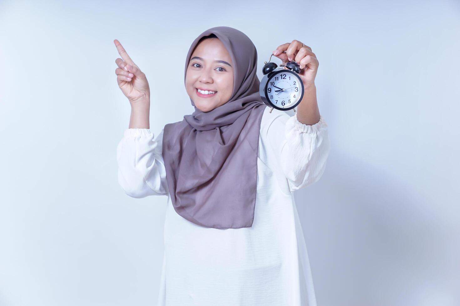 Smiling asian woman in a hijab holds an alarm clock pointing upward isolated on white background photo