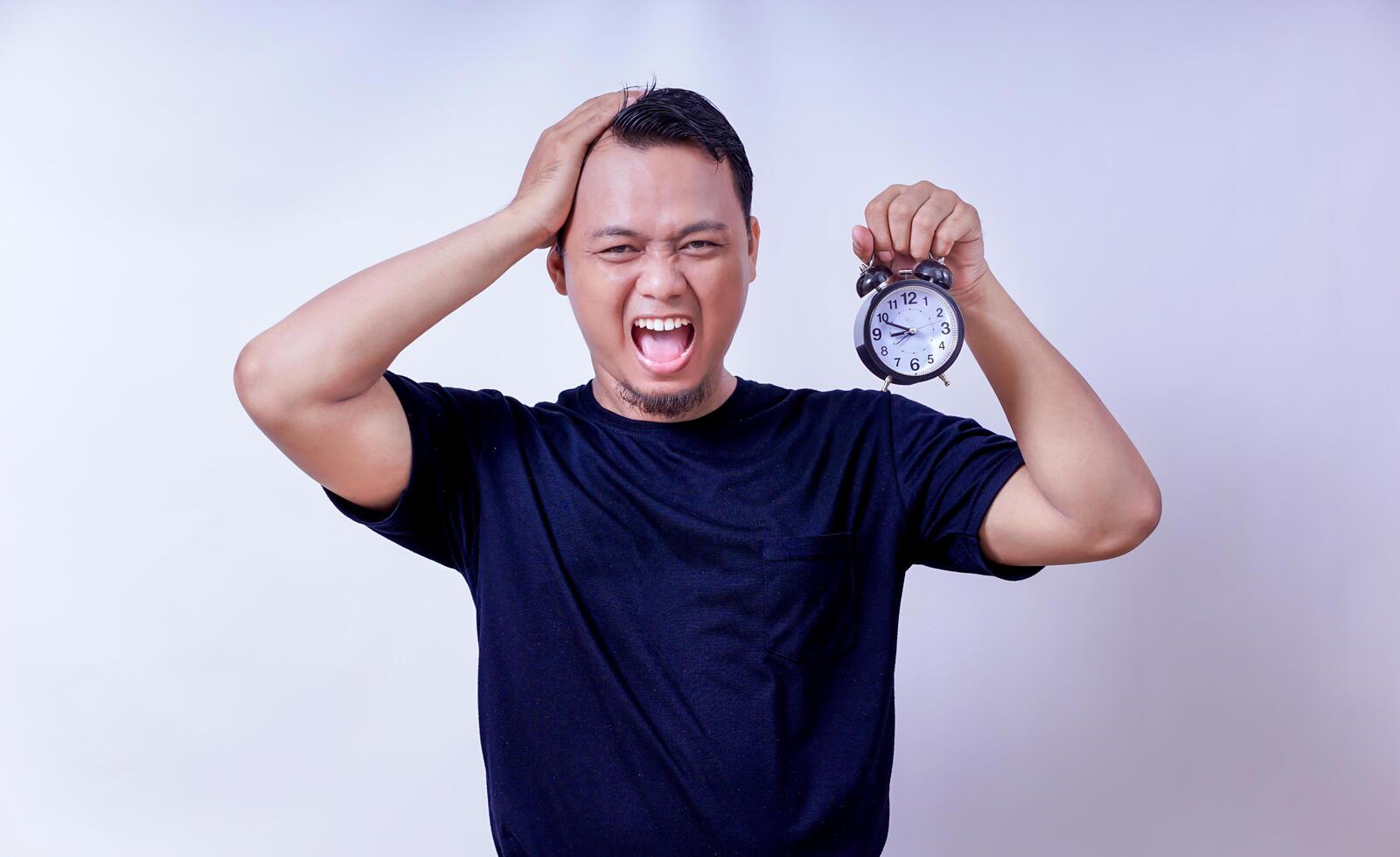 An asian man looks frustrated while holding an alarm clock, emphasizing time pressure and stress isolated on white background photo