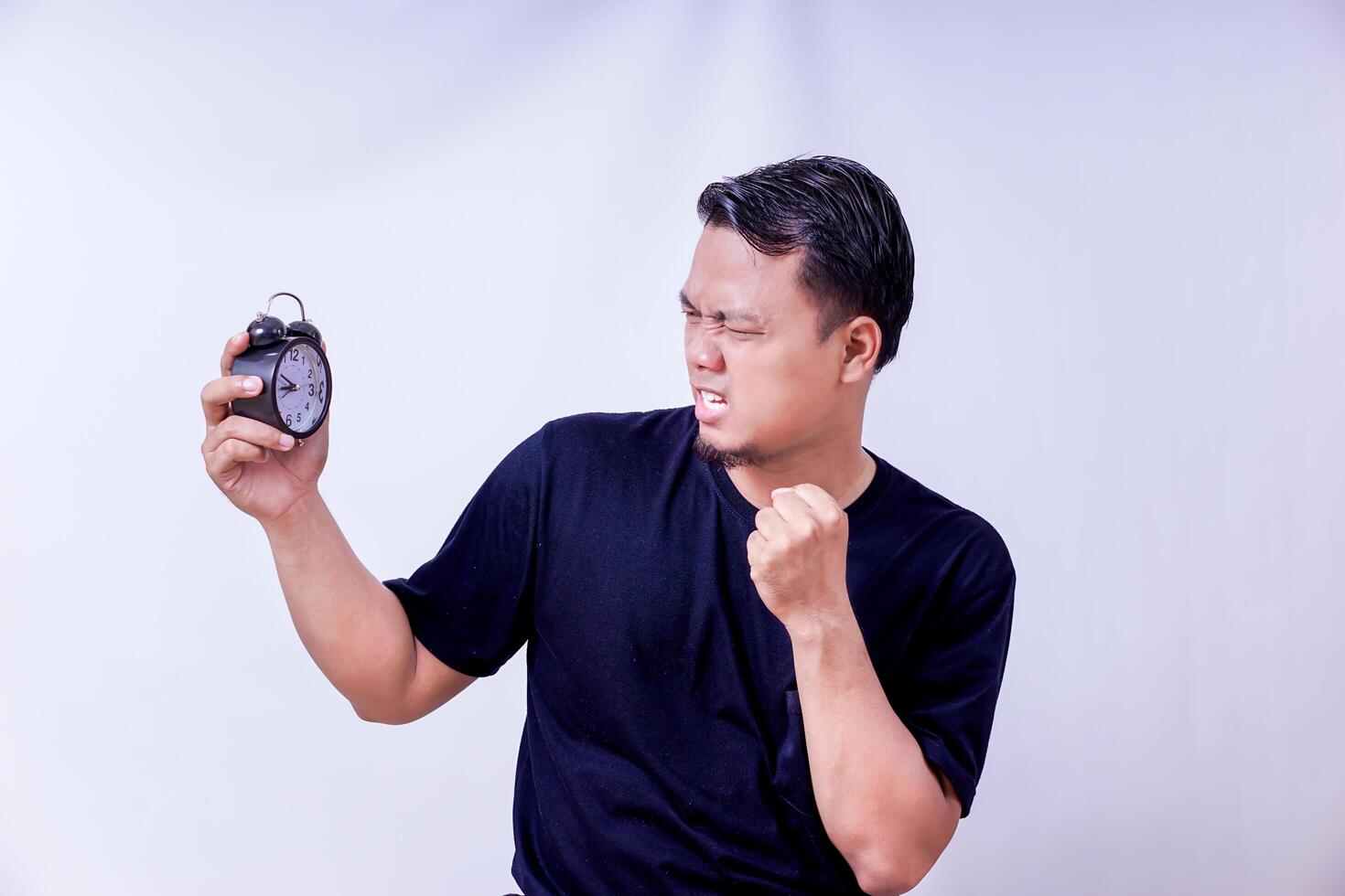 A frustrated asian man checks an alarm clock, expressing urgency or annoyance at the time isolated on white background photo