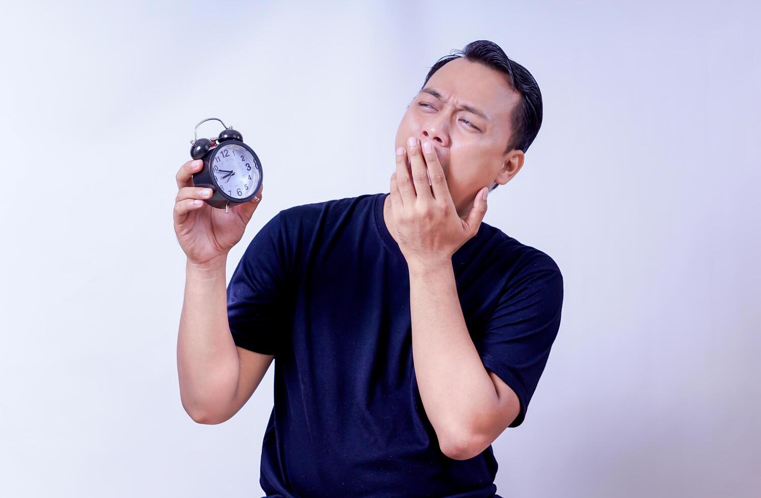 An asian man yawning while holding an alarm clock, expressing surprise isolated on white background photo