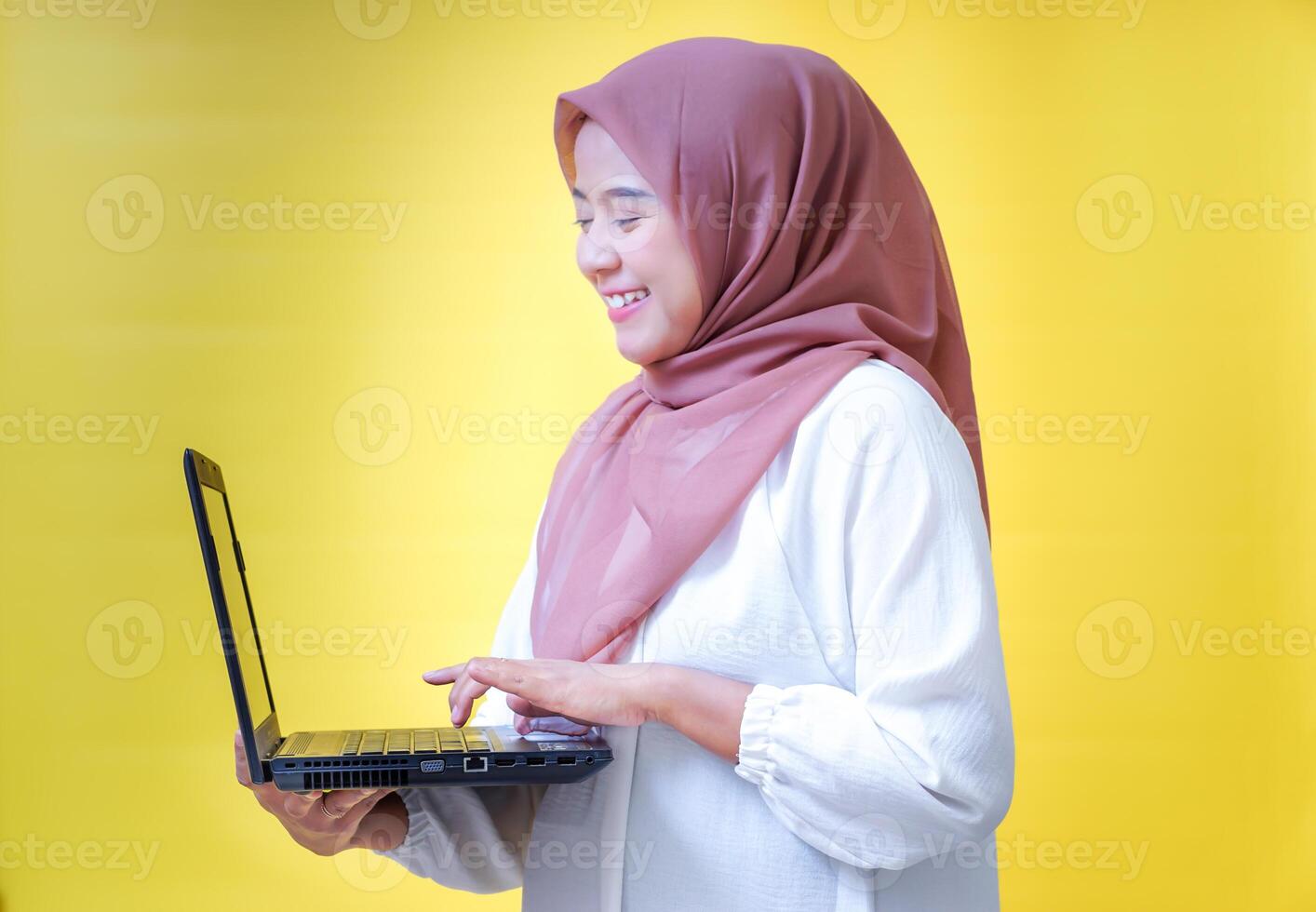 Smiling young asian woman in a white outfit and hijab smiles while holding a laptop against a yellow background photo
