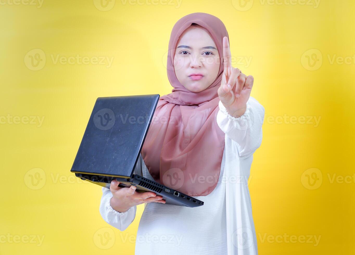 An asian woman in a hijab holds a laptop, raising her finger to emphasize an important point. photo