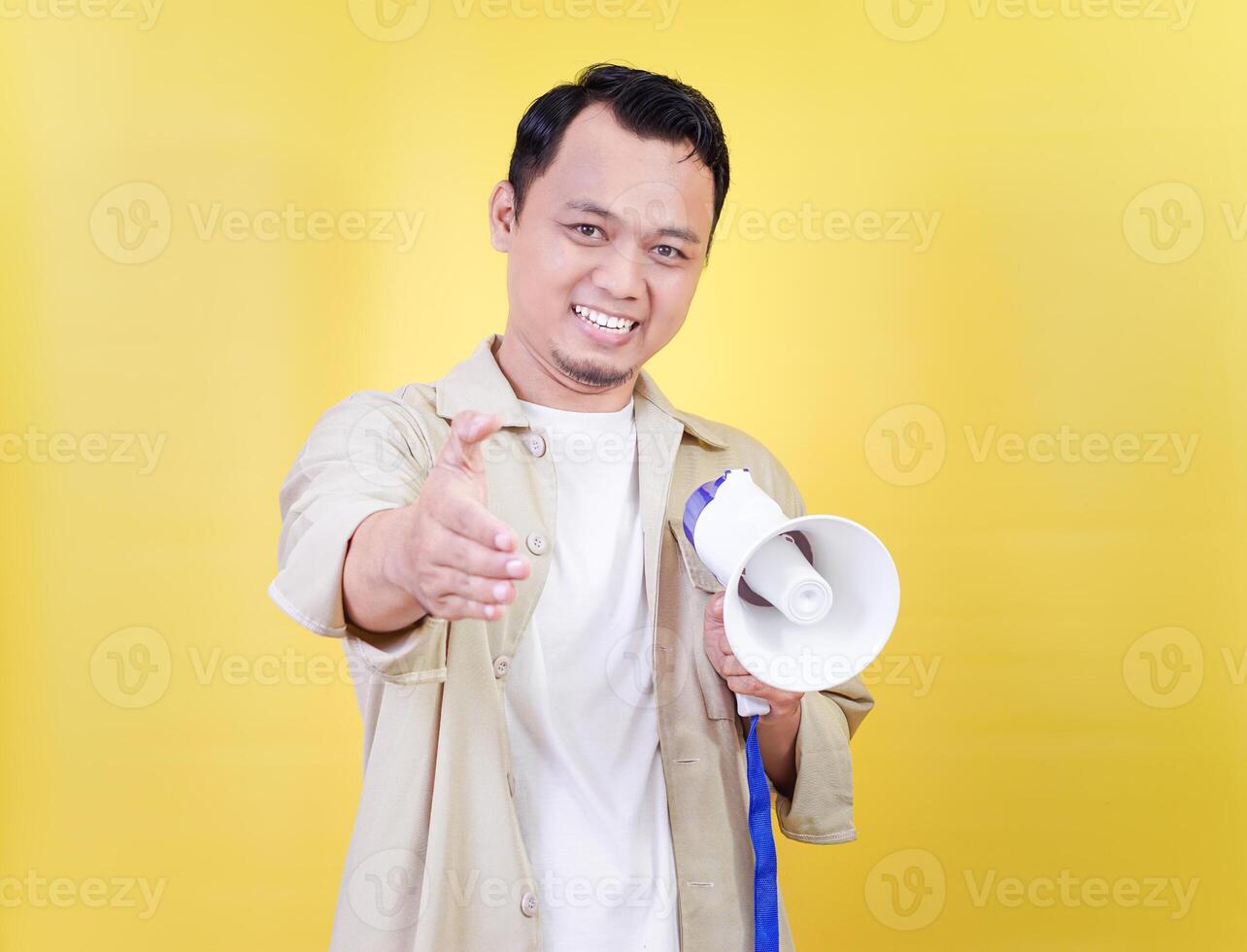 A smiling asian man in a light jacket extends his hand, holding a megaphone against a yellow background. photo