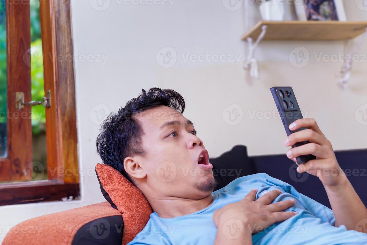 A surprised asian man reacts while watching something on his phone, capturing a moment of shock. photo