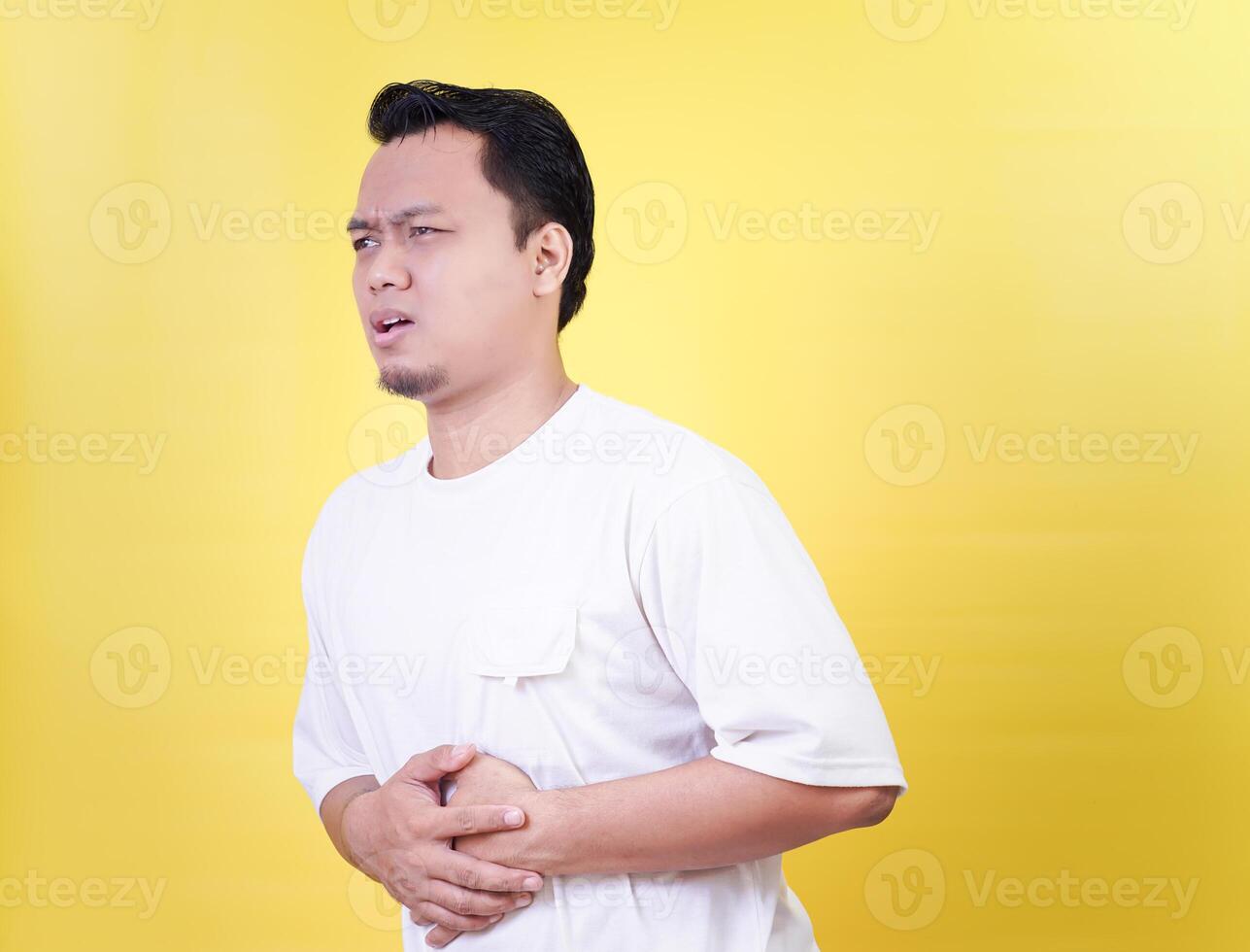 An asian man in white shirt appears to be in discomfort, holding his stomach against a yellow background photo
