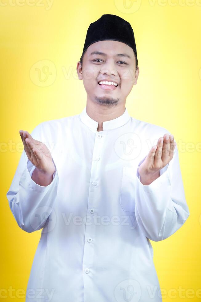 A smiling asian young man in a white outfit welcomes with open hands against a bright yellow background photo
