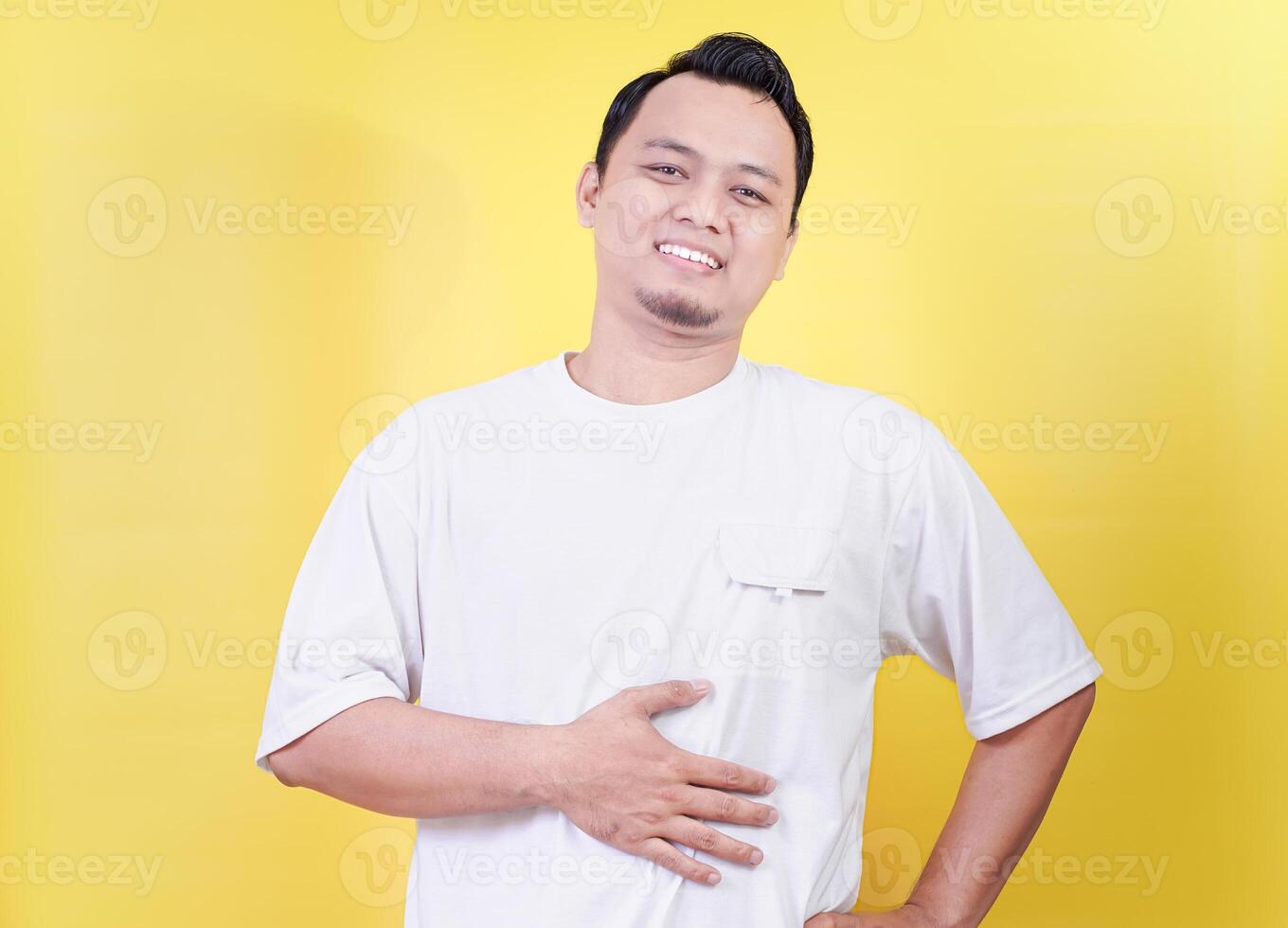 A smiling asian man in a white shirt poses confidently while holding his stomach against a yellow background photo