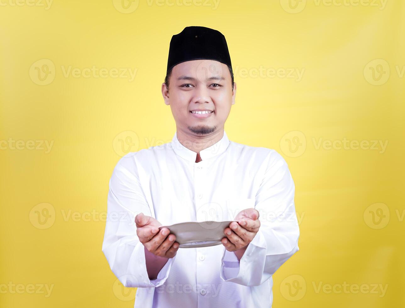 A smiling asian muslim man in traditional attire holding a plate with a yellow background photo