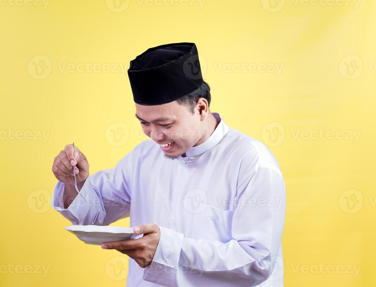 An asian muslim man happily enjoying a meal, wearing traditional attire against a vibrant yellow background. photo