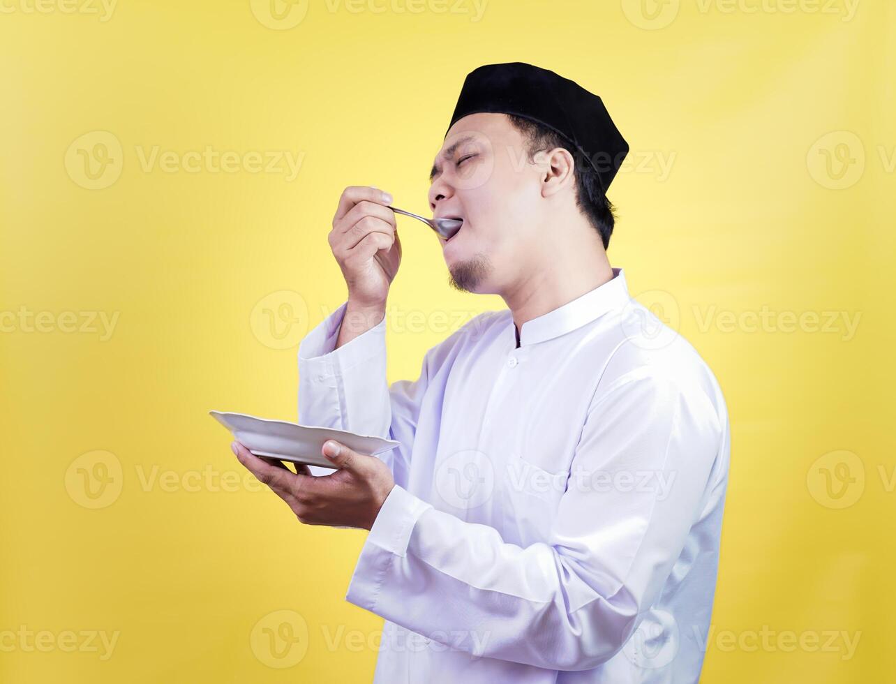 asian muslim man in traditional attire enjoys tasty food with a satisfied expression against a yellow background photo