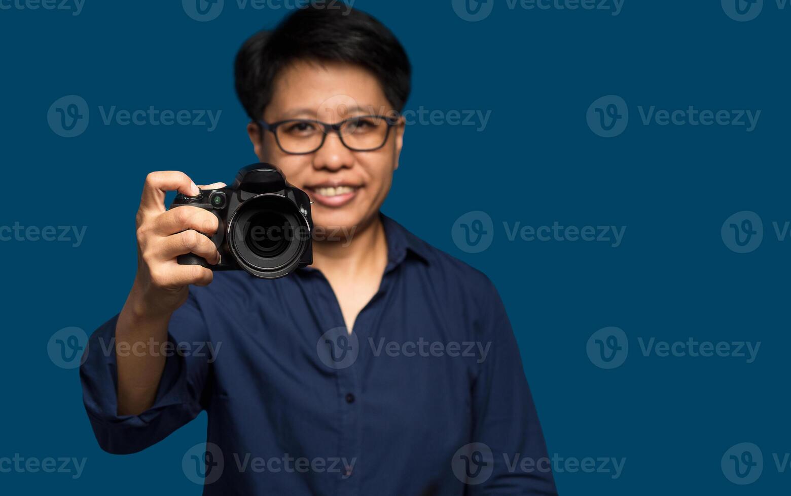A woman photographer with short black hair holds a digital camera while standing on a blue background photo