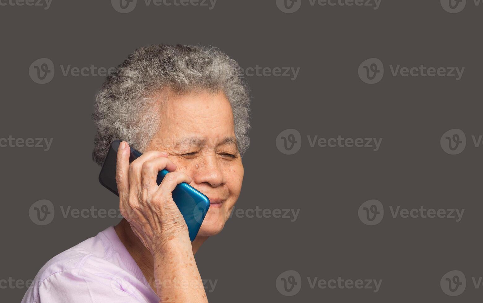 An elderly Asian woman is talking on a smartphone with a smile while standing on a gray background photo