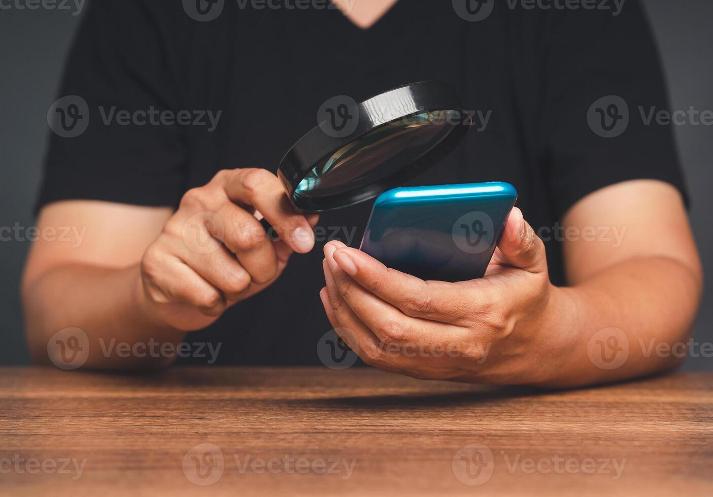 A young man in a black casual is holding a magnifying glass and looking at a smartphone while sitting at the table photo