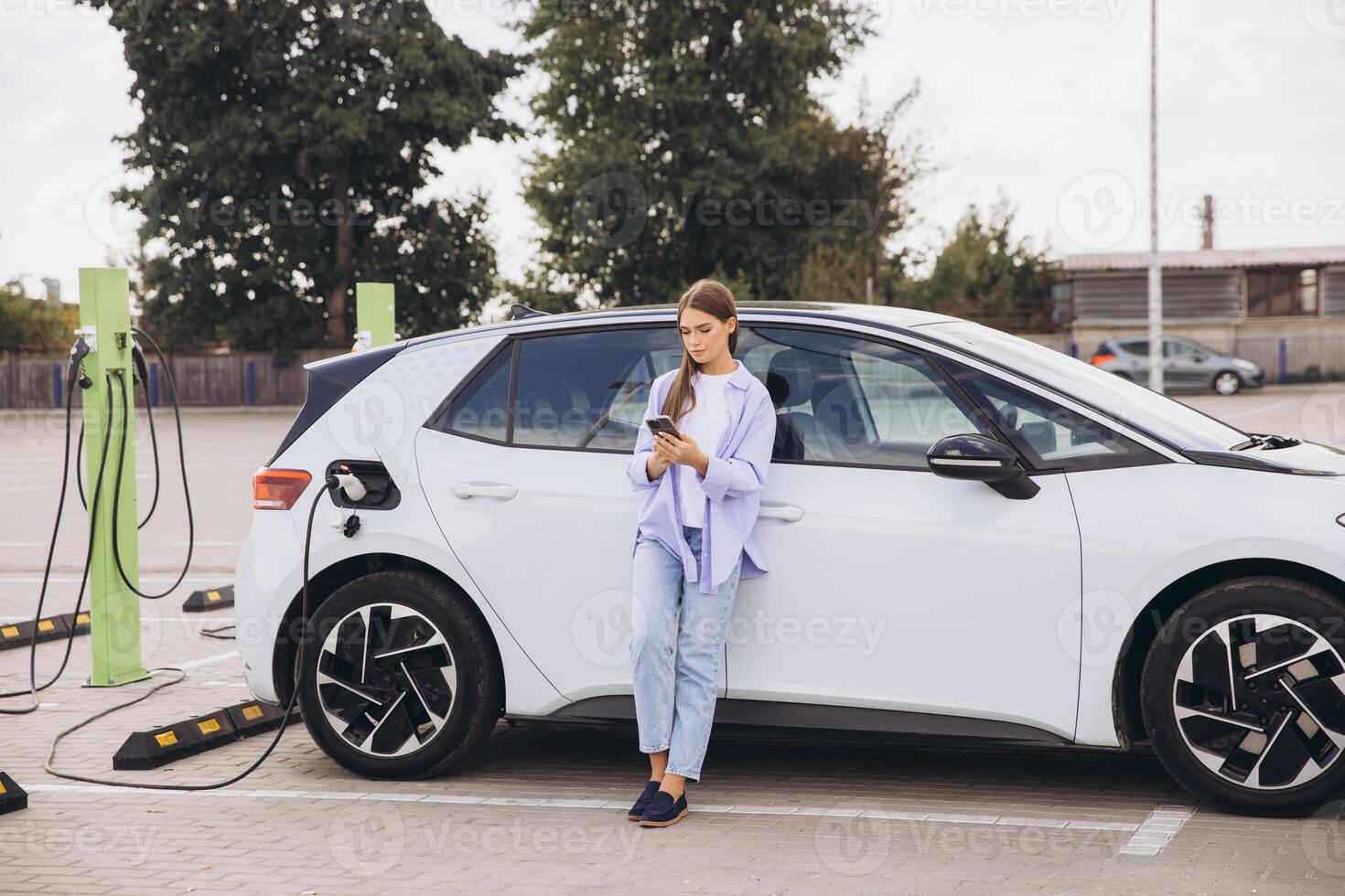 Young Woman Charging Electric Car at Urban Station While Using Smartphone for Eco-Friendly Travel and Sustainable Energy Lifestyle photo
