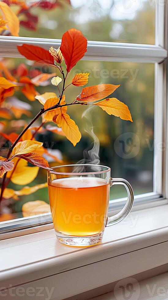 A steaming glass cup of hot tea on a windowsill, with vibrant autumn leaves outside the window, creating a warm and cozy fall atmosphere, steaming cup of apple cider photo