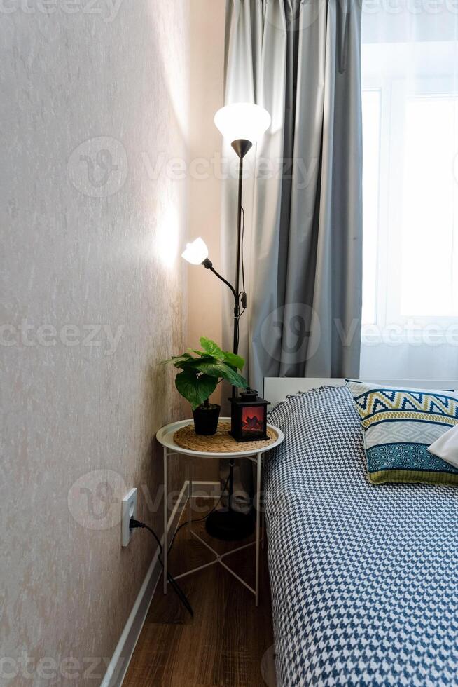 This is a cozy bedroom that features a comfortable bed, a stylish nightstand alongside it, a soft lamp providing warm light, and a window allowing natural sunlight to brighten the space photo