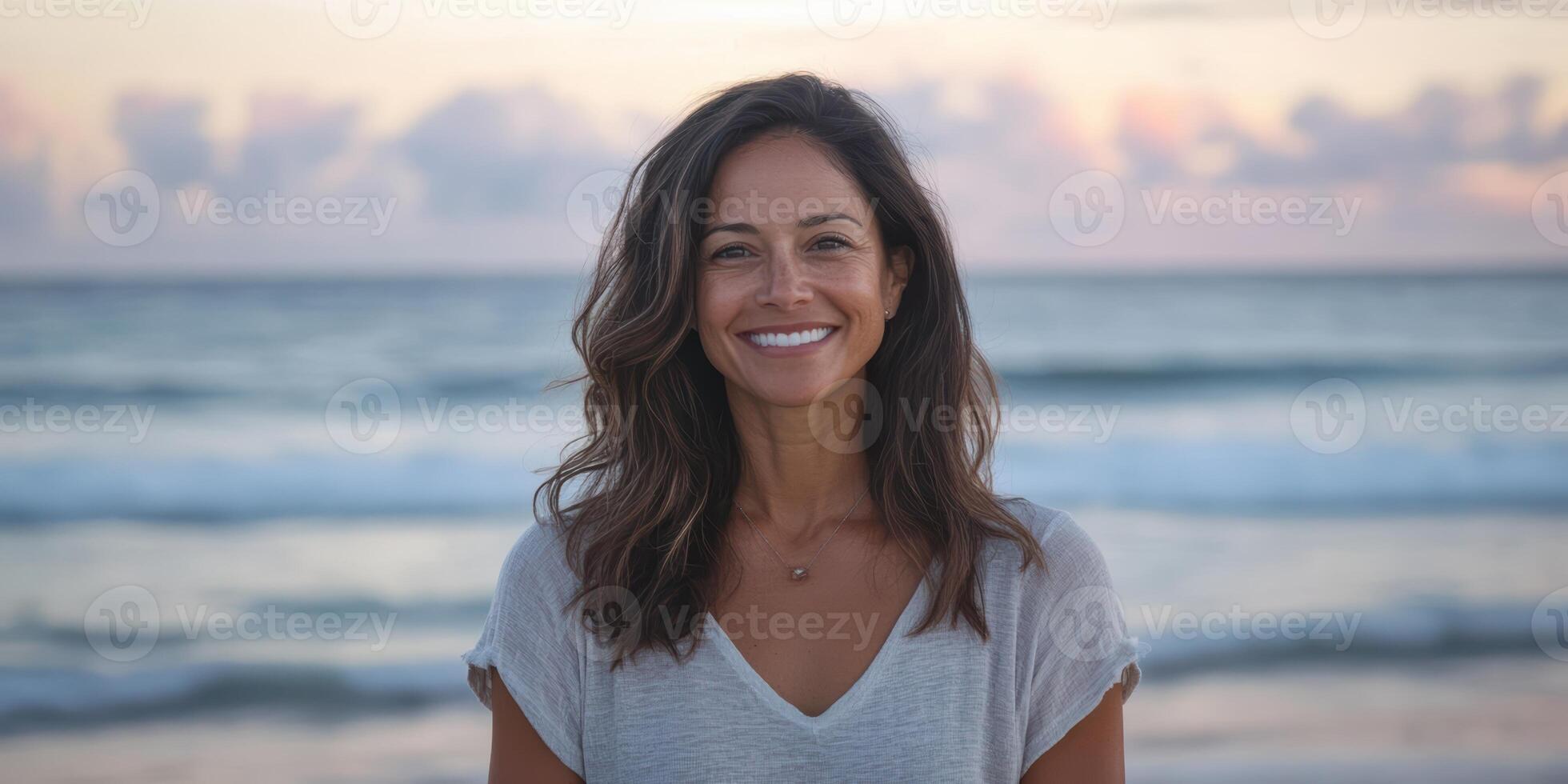 A woman with long brown hair is smiling at the camera on a beach. She is wearing a white shirt and a necklace. Scene is happy and relaxed, as the woman is enjoying her time at the beach photo