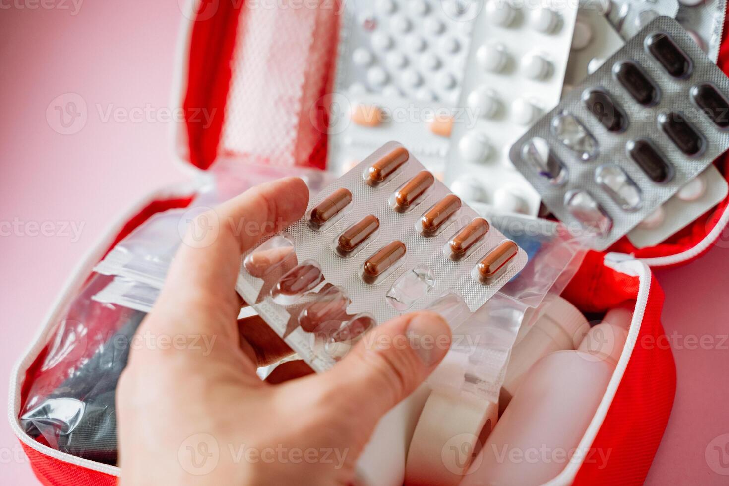A person is currently holding a blister pack that contains various pills right in front of a first aid kit that is wellstocked with medical supplies for emergencies and health care needs photo