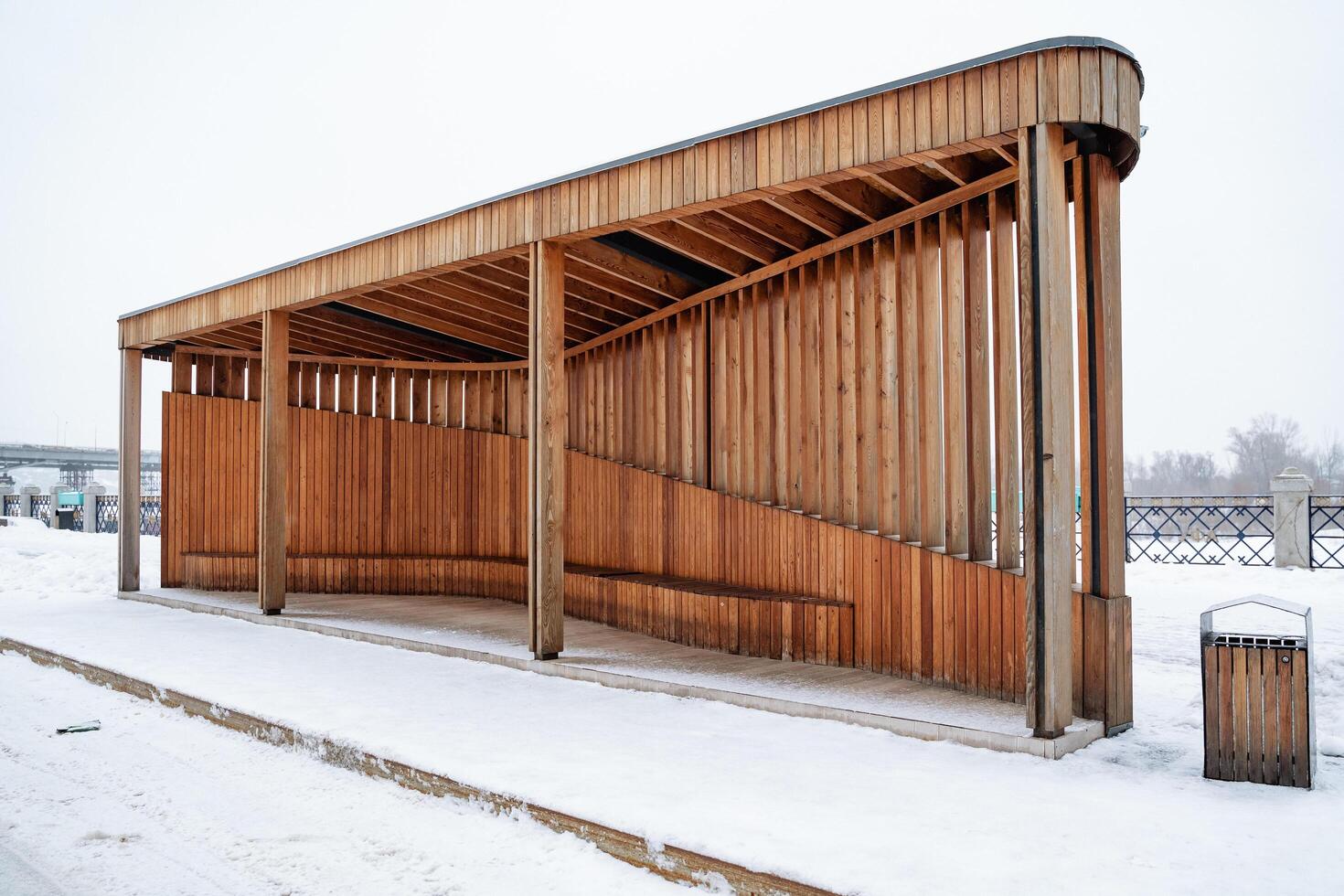 In a serene winter landscape, a charming wooden bus stop stands amid glistening snow, complete with a sturdy bench for waiting passengers and a trash can for cleanliness photo