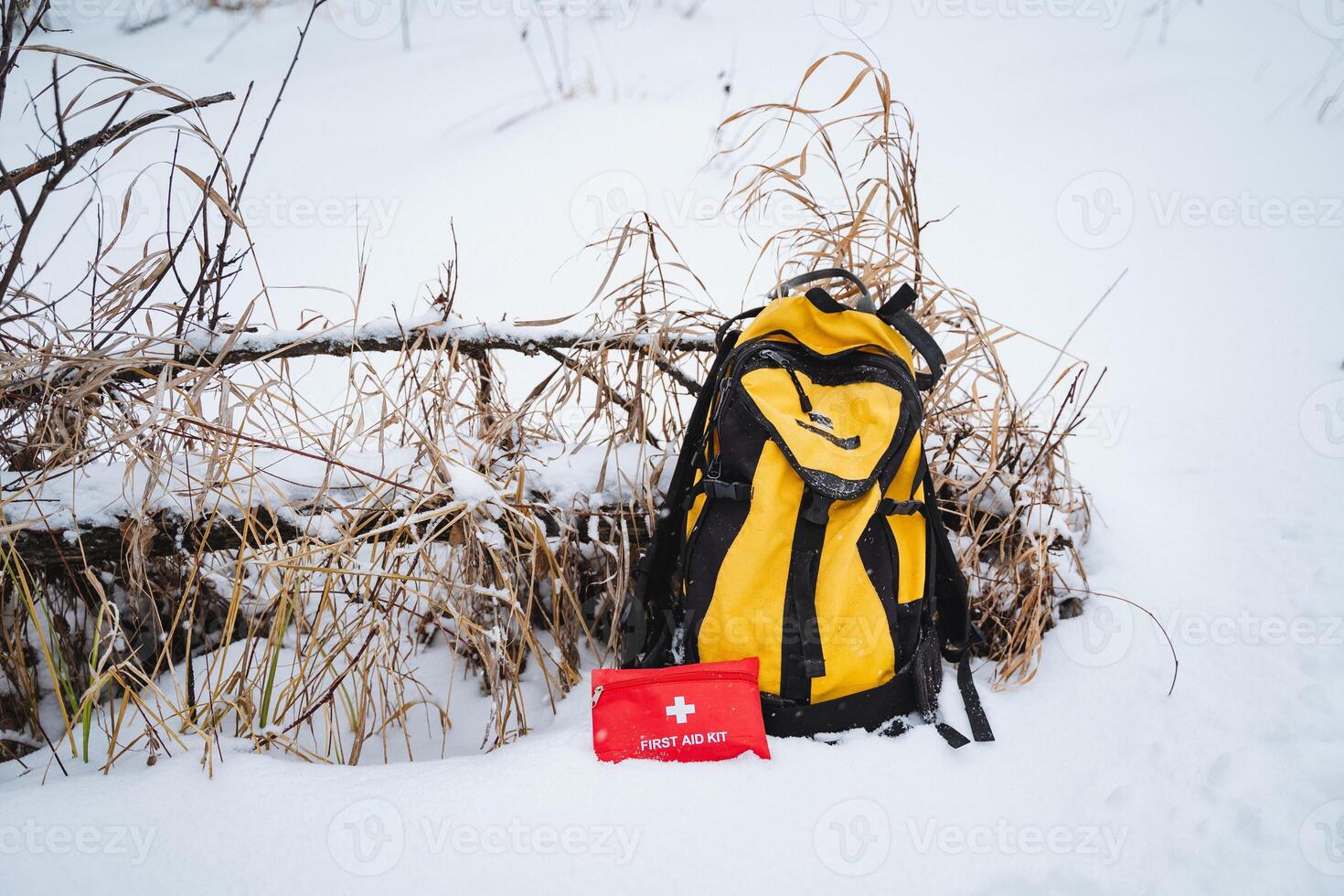 In the winter landscape, a bright yellow backpack is resting in the soft, fluffy, white snow, positioned right beside a vibrant red first aid kit, creating a beautifully striking contrast photo