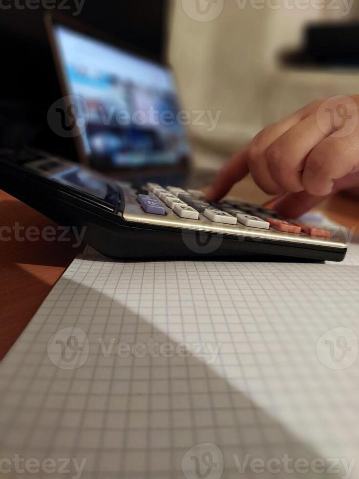 Hand using calculator on graph paper desk with open laptop in background photo