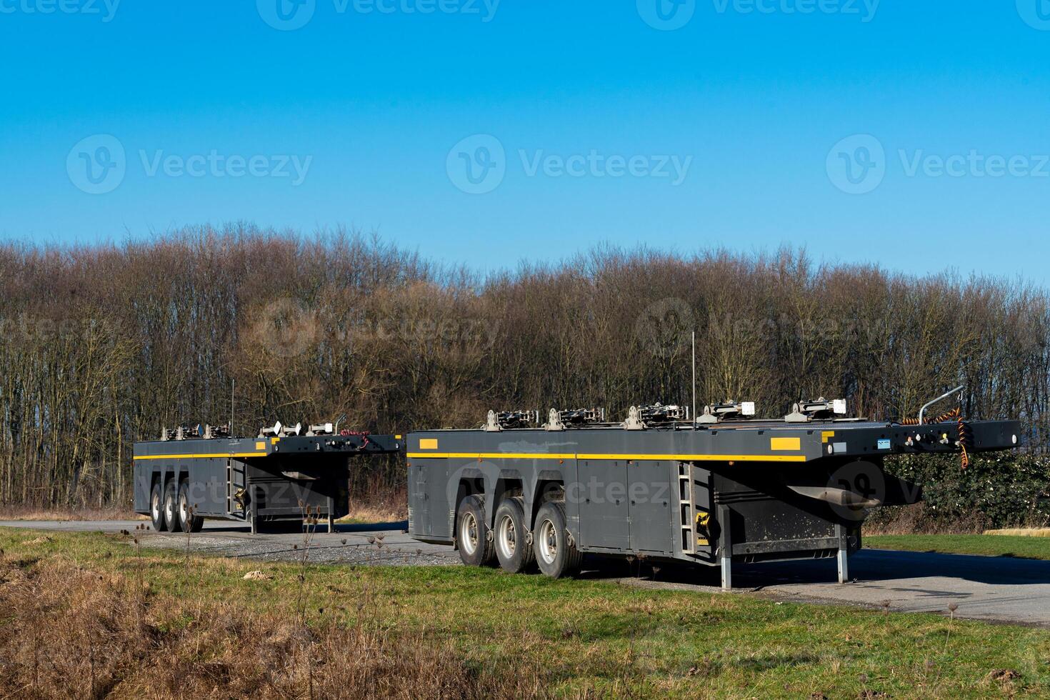 Two heavy machinery trailers parked by a wooded area on a sunny day photo