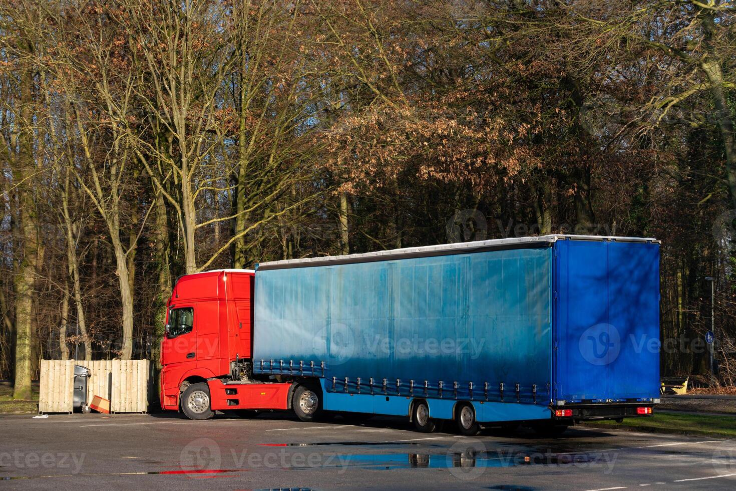 A large red truck with a blue trailer is parked in a forest area. It photo
