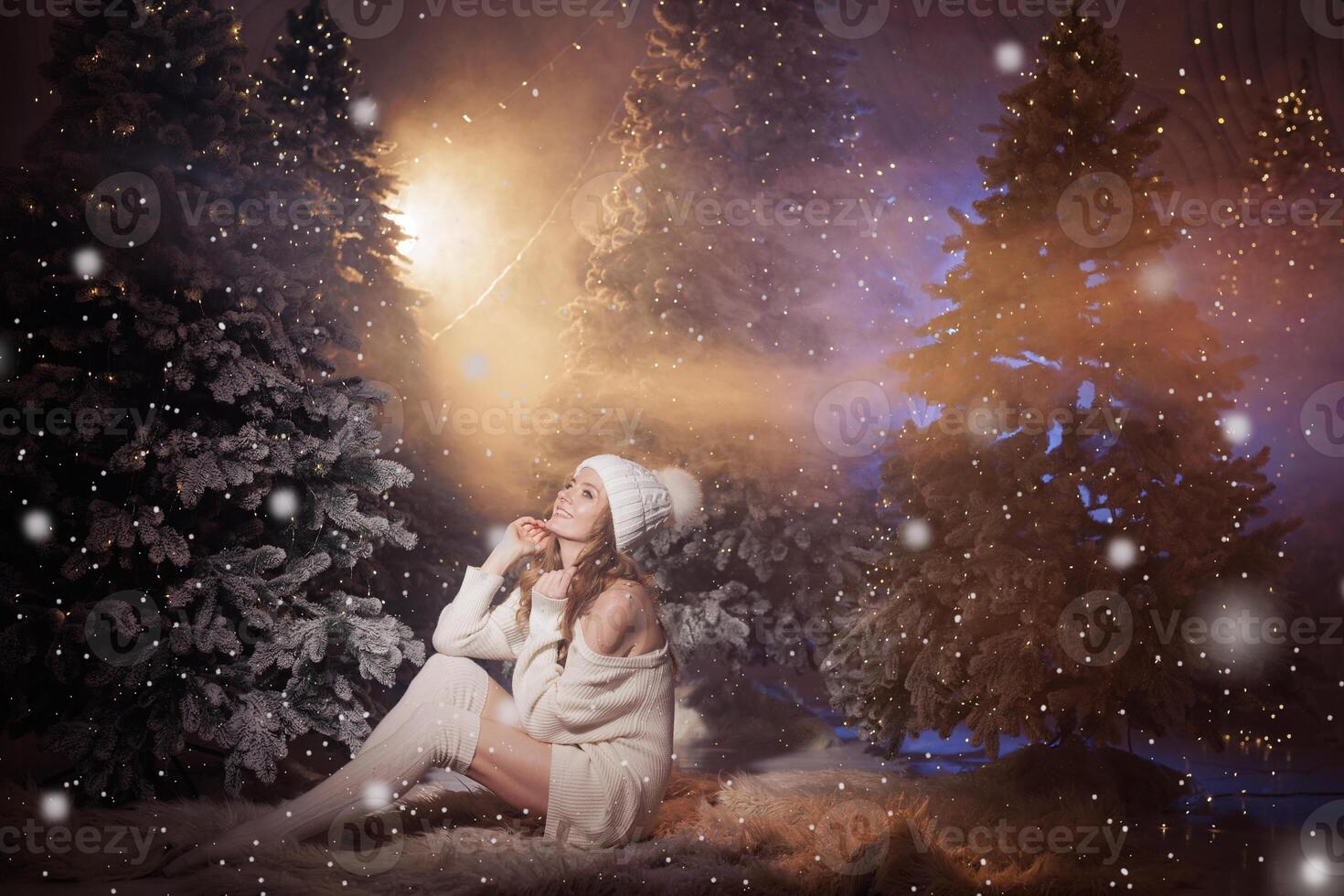 A woman in a cozy white sweater sits in a winter snowy forest and enjoys the flying snow, the magical winter atmosphere photo