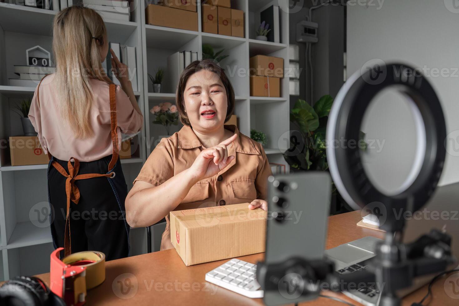 E-commerce entrepreneur explaining shipment process on camera with package, while assistant organizes inventory photo