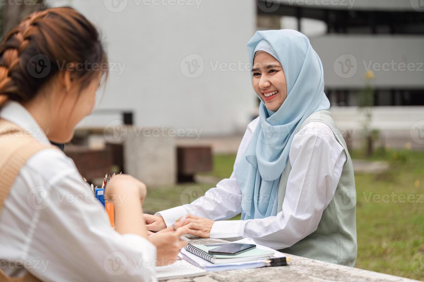 Female student in hijab smiling while studying with a friend outdoors. photo