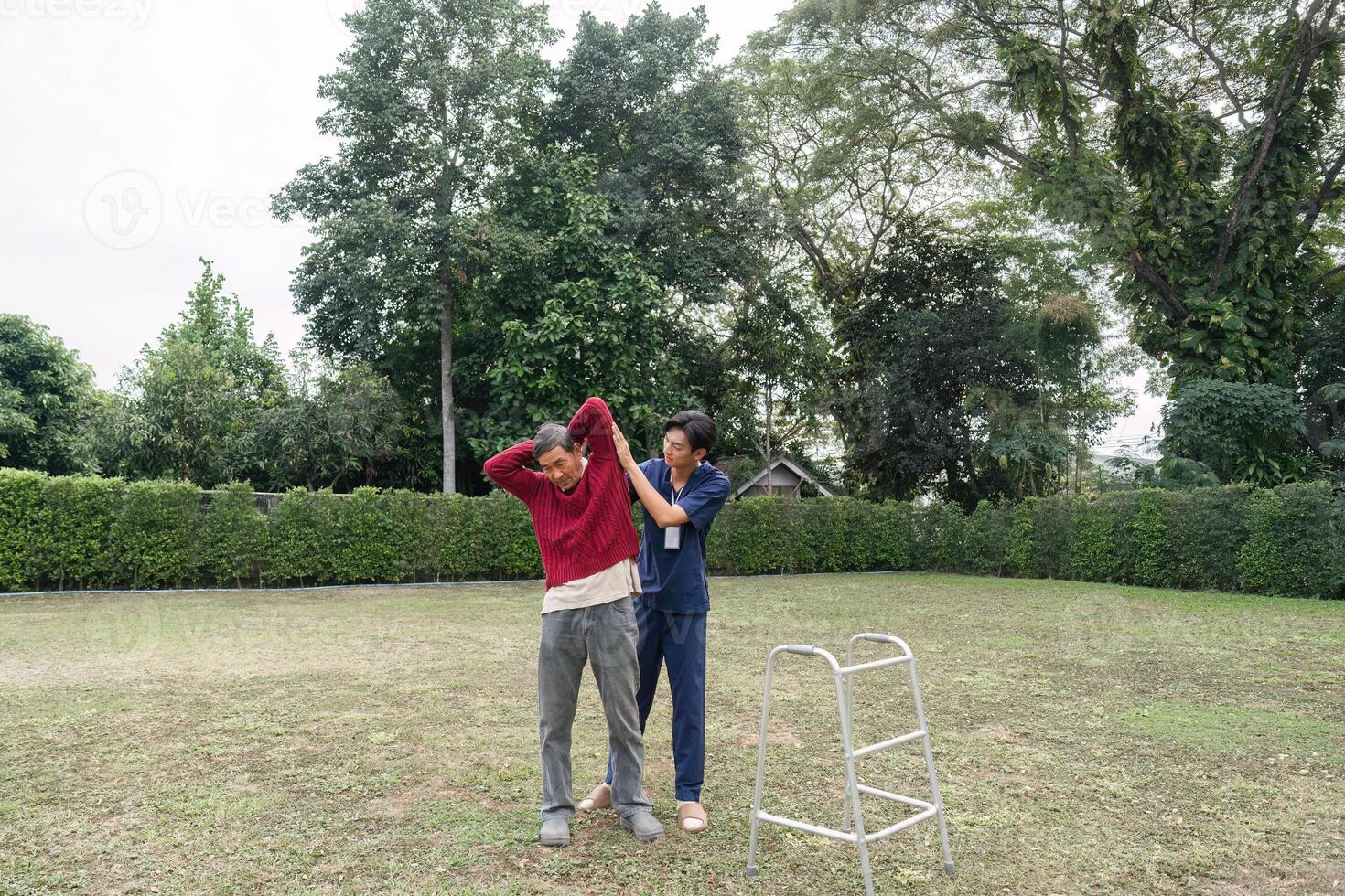 Young caregiver supporting elderly man with stretching exercise using walker in garden photo