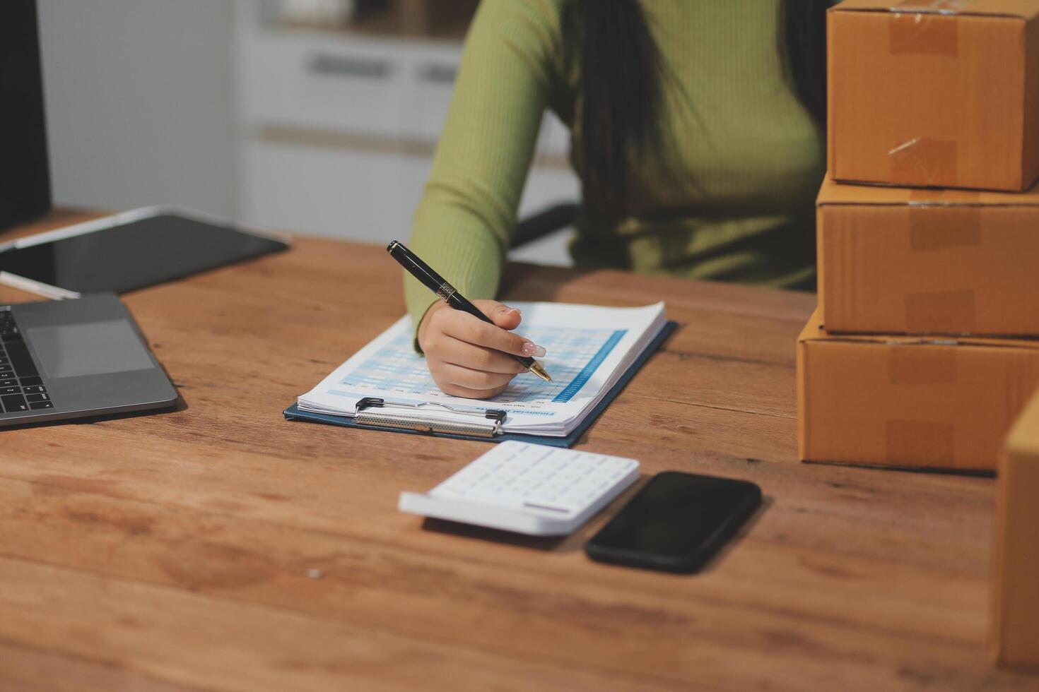 A woman is writing on a notebook while sitting at a desk with boxes and a laptop photo