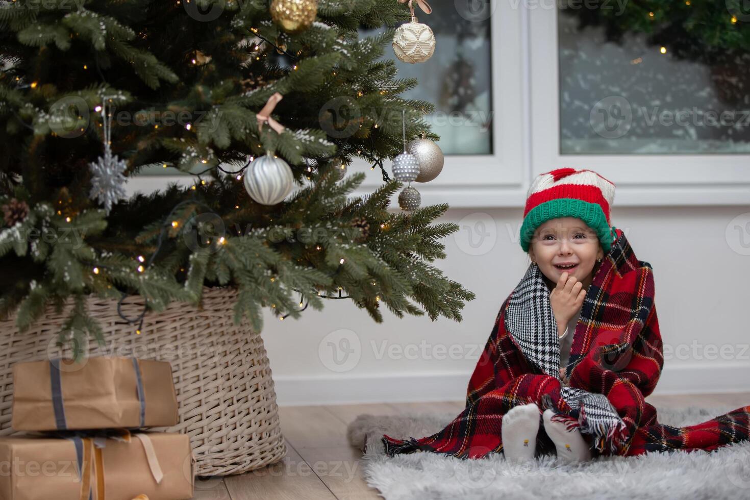 A happy little girl in a Santa Claus hat is wrapped in a warm plaid blanket by the Christmas tree photo