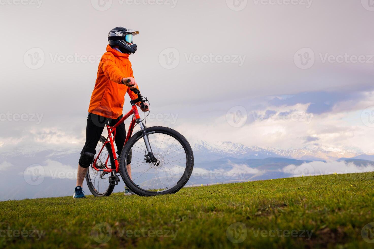 man rides a mountain bike on top of a green hill. A guy in a protective helmet stands on a meadow looking into the distance, with large snow-capped mountains in the background among clouds photo