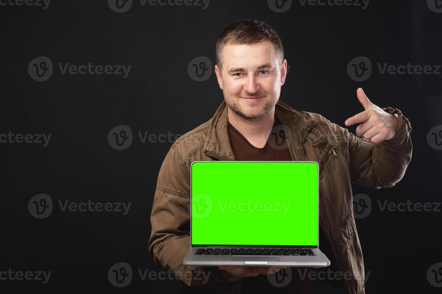 Young male IT specialist in leather jacket holding work on blank laptop screen isolated on plain green background. Smiling man gesturing at computer, place for your ad photo
