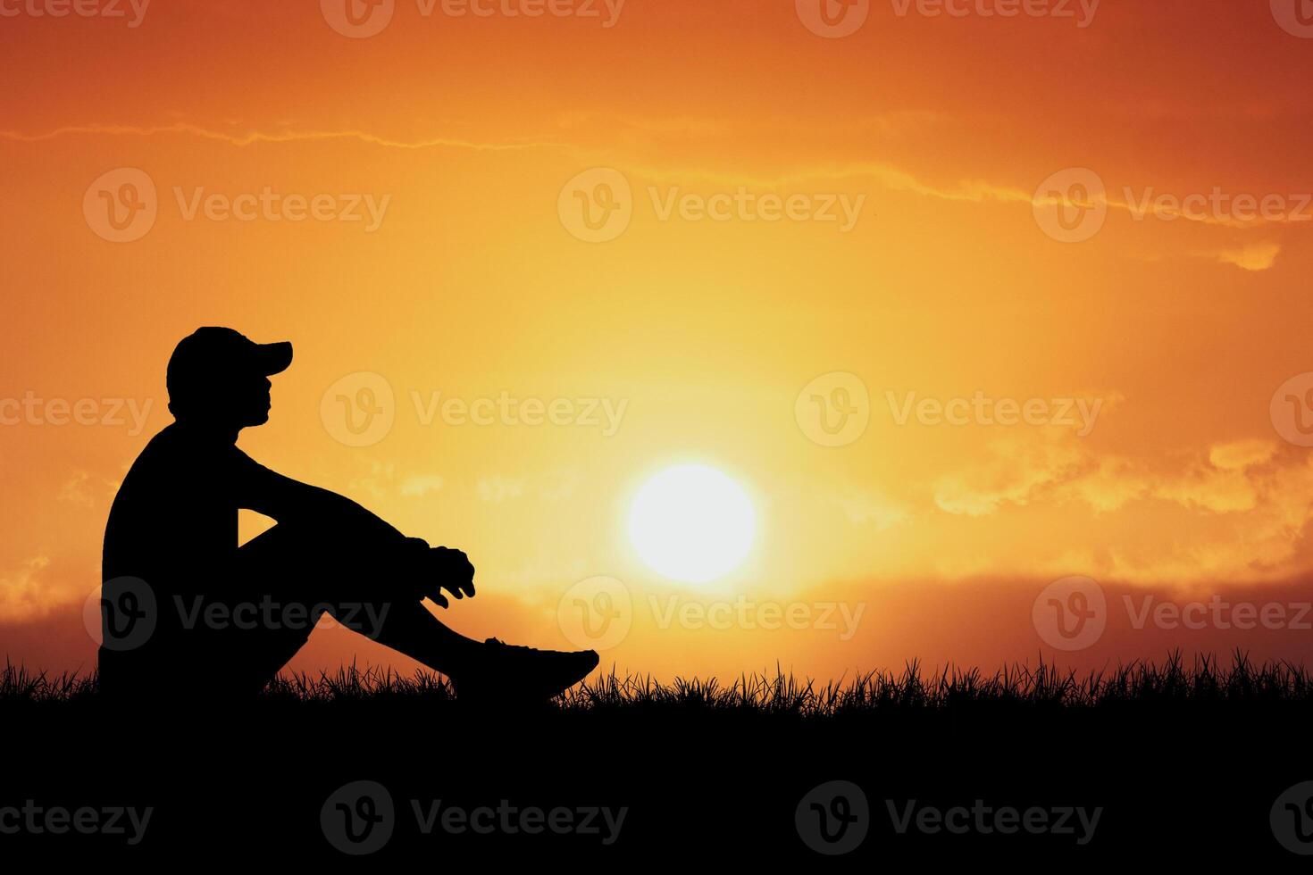 Silhouette of a Person Sitting on a Hill at Sunset with Vibrant Orange Sky and Sun in the Background photo