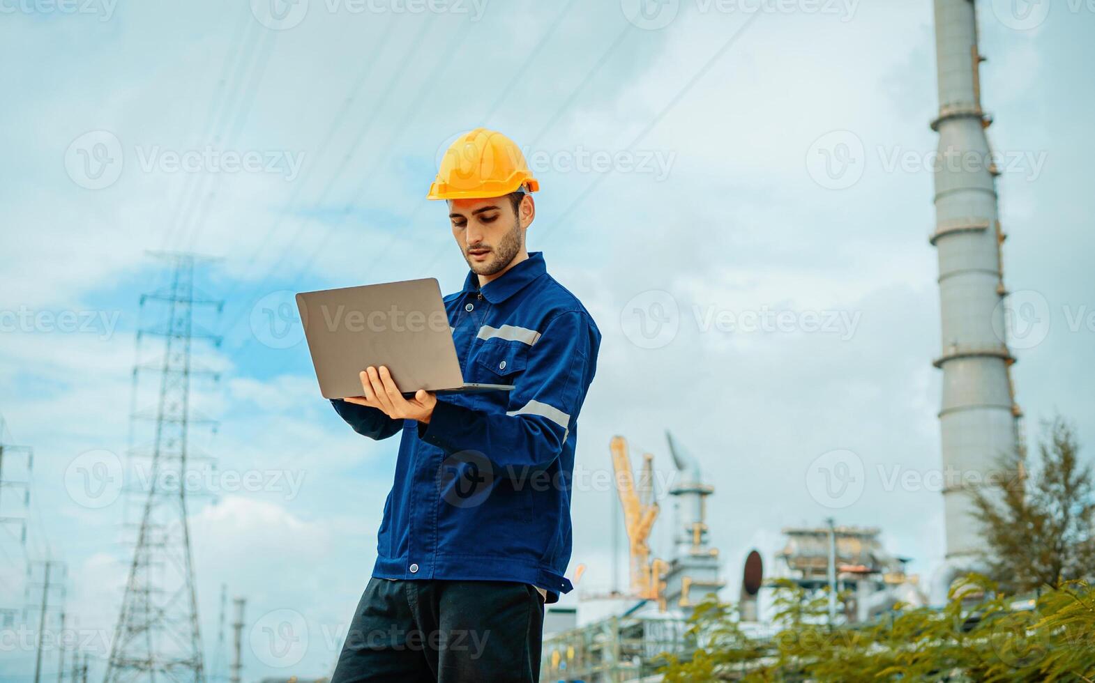 Worker in safety gear using laptop at industrial site with power lines in the background during the day photo