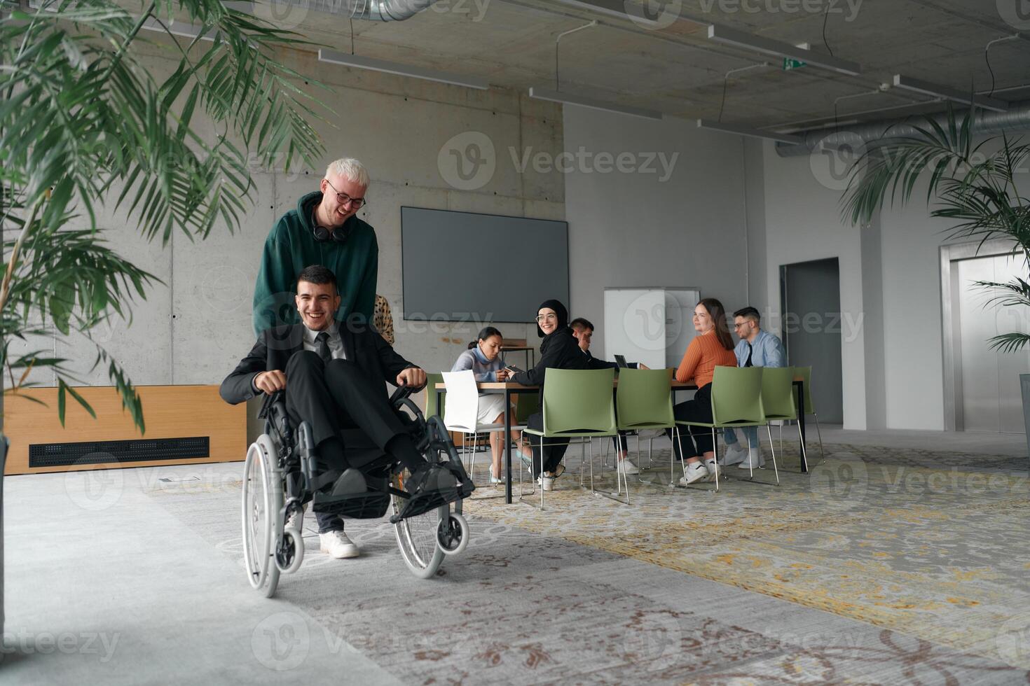 A diverse and multicultural business team collaborates in the office while their colleagues, including a director in a wheelchair and his friend, share a lighthearted moment, fostering an atmosphere photo