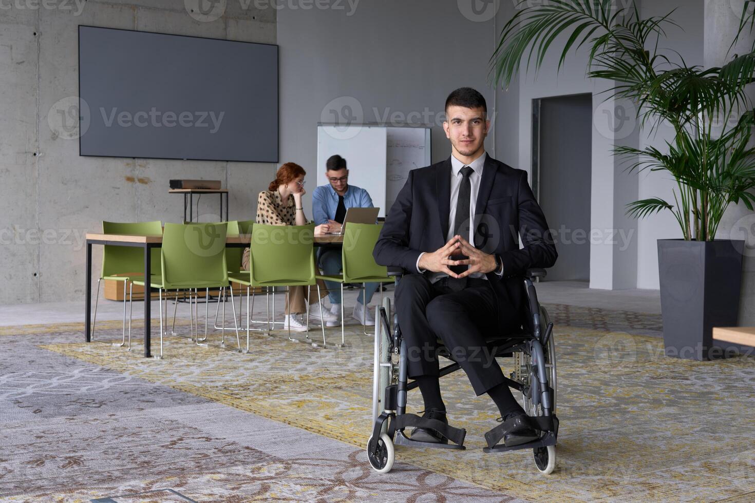 Business Director in a Suit Using a Wheelchair with Colleagues Conducting a Meeting Behind Him photo