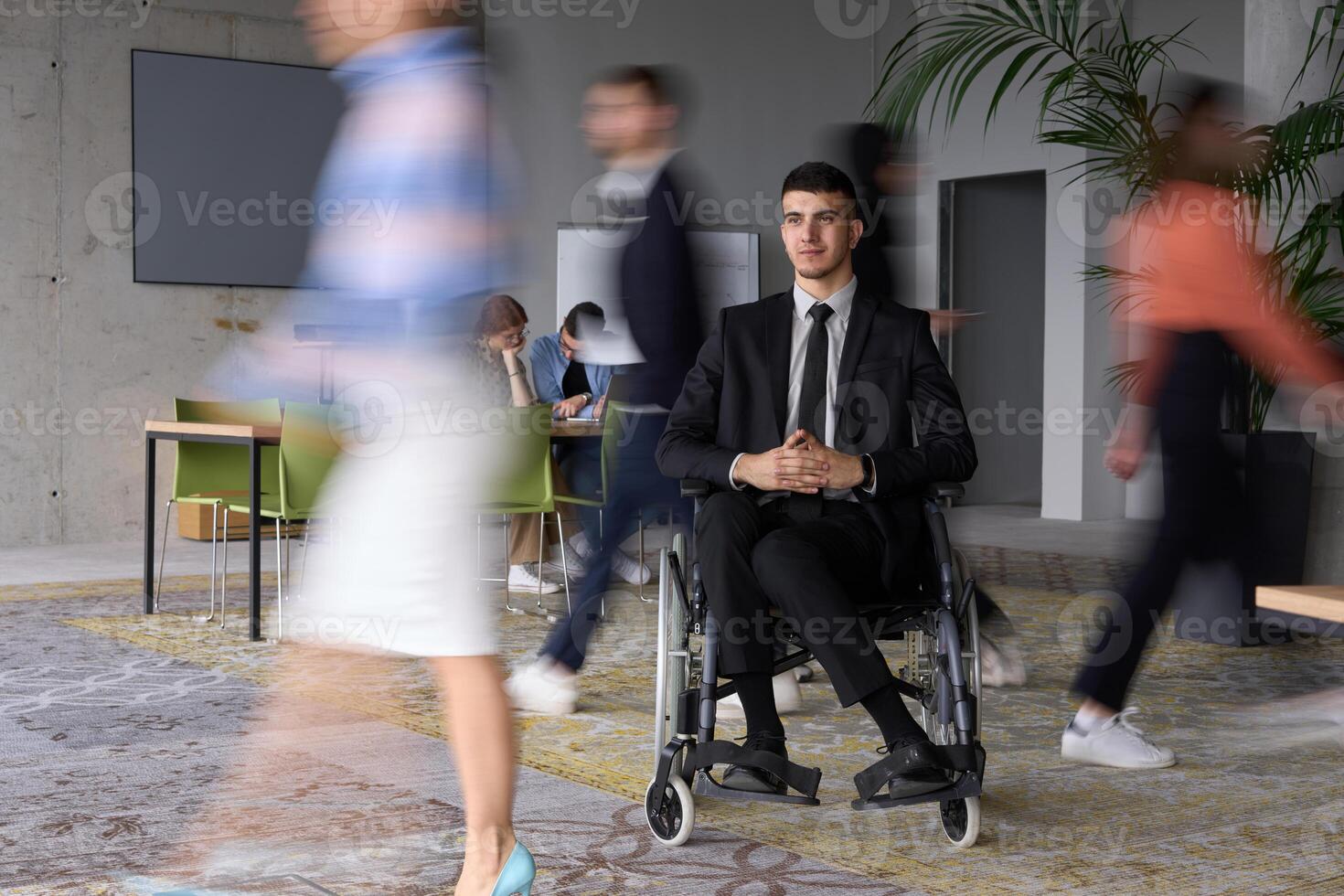 A businessman in a wheelchair navigating through a busy office, surrounded by his colleagues who are actively engaged in their work and collaboration photo