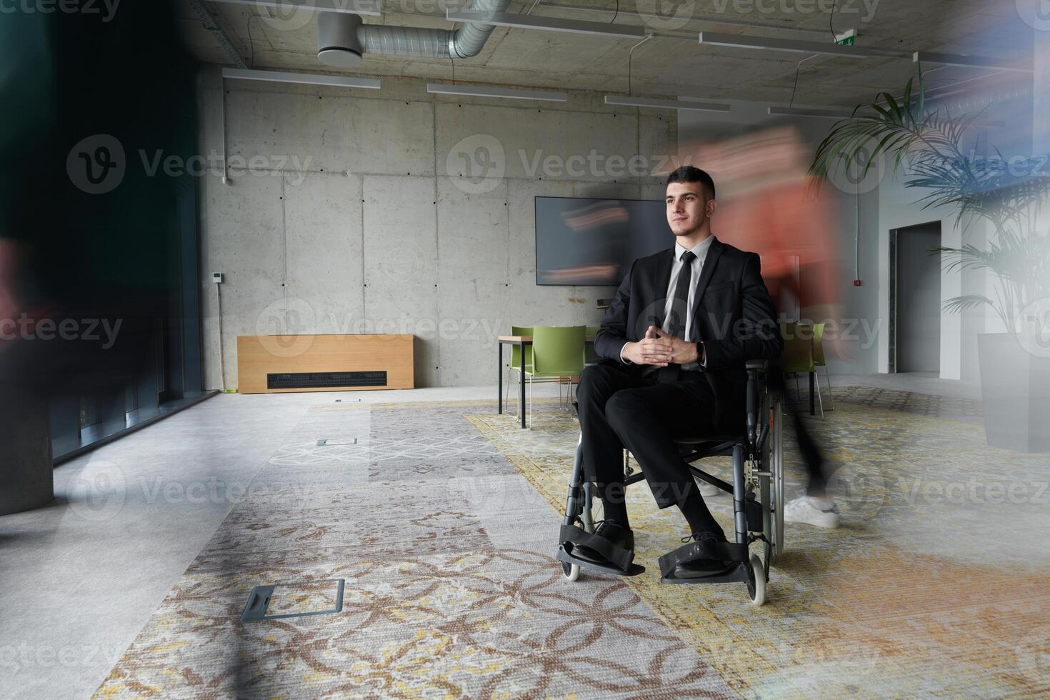 A businessman in a wheelchair navigating through a busy office, surrounded by his colleagues who are actively engaged in their work and collaboration photo
