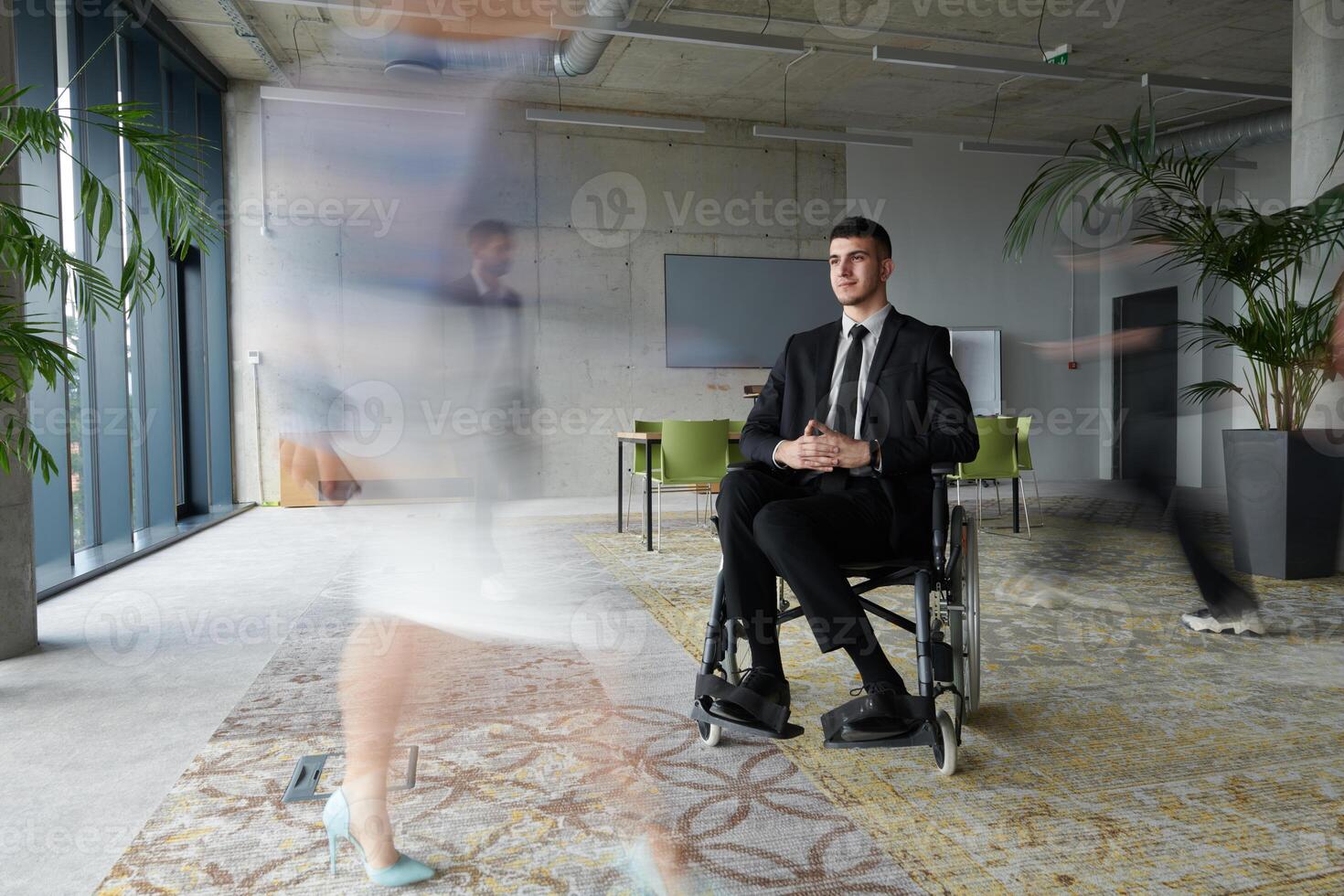 A businessman in a wheelchair navigating through a busy office, surrounded by his colleagues who are actively engaged in their work and collaboration photo