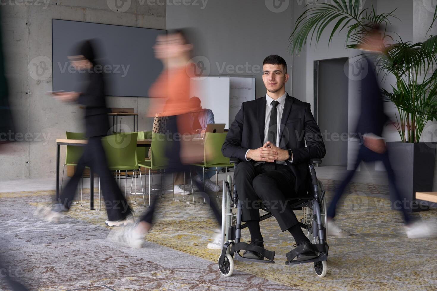 A businessman in a wheelchair navigating through a busy office, surrounded by his colleagues who are actively engaged in their work and collaboration photo