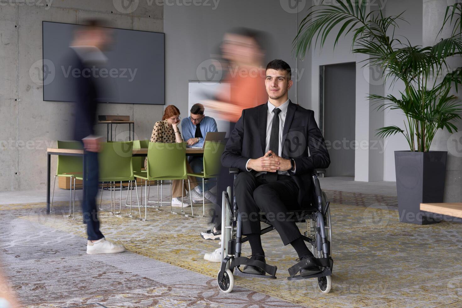 A businessman in a wheelchair navigating through a busy office, surrounded by his colleagues who are actively engaged in their work and collaboration photo