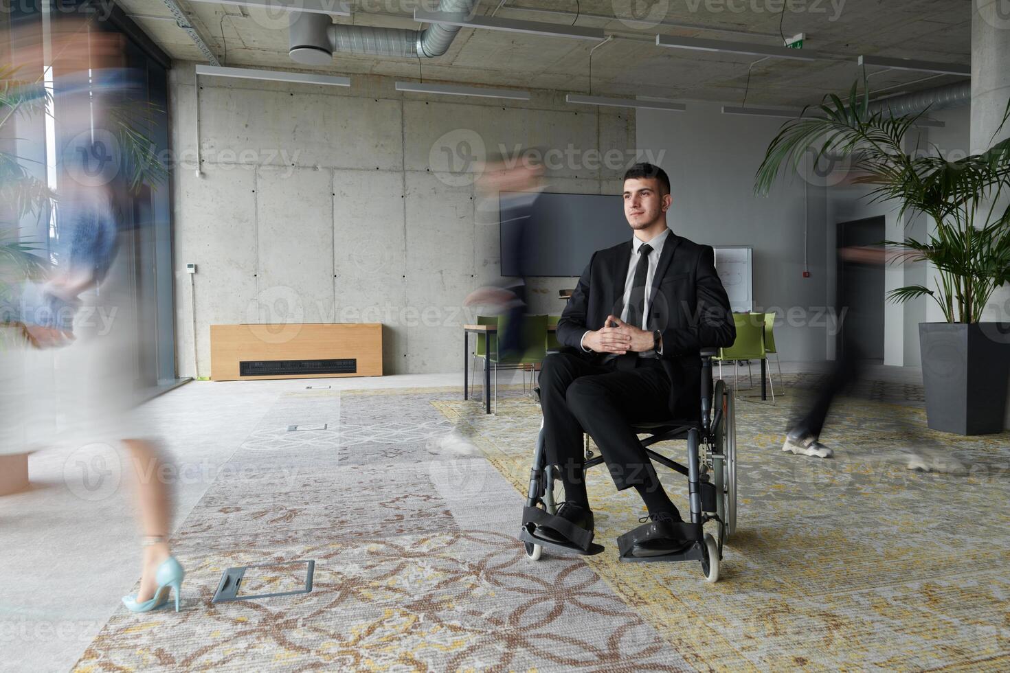 A businessman in a wheelchair navigating through a busy office, surrounded by his colleagues who are actively engaged in their work and collaboration photo
